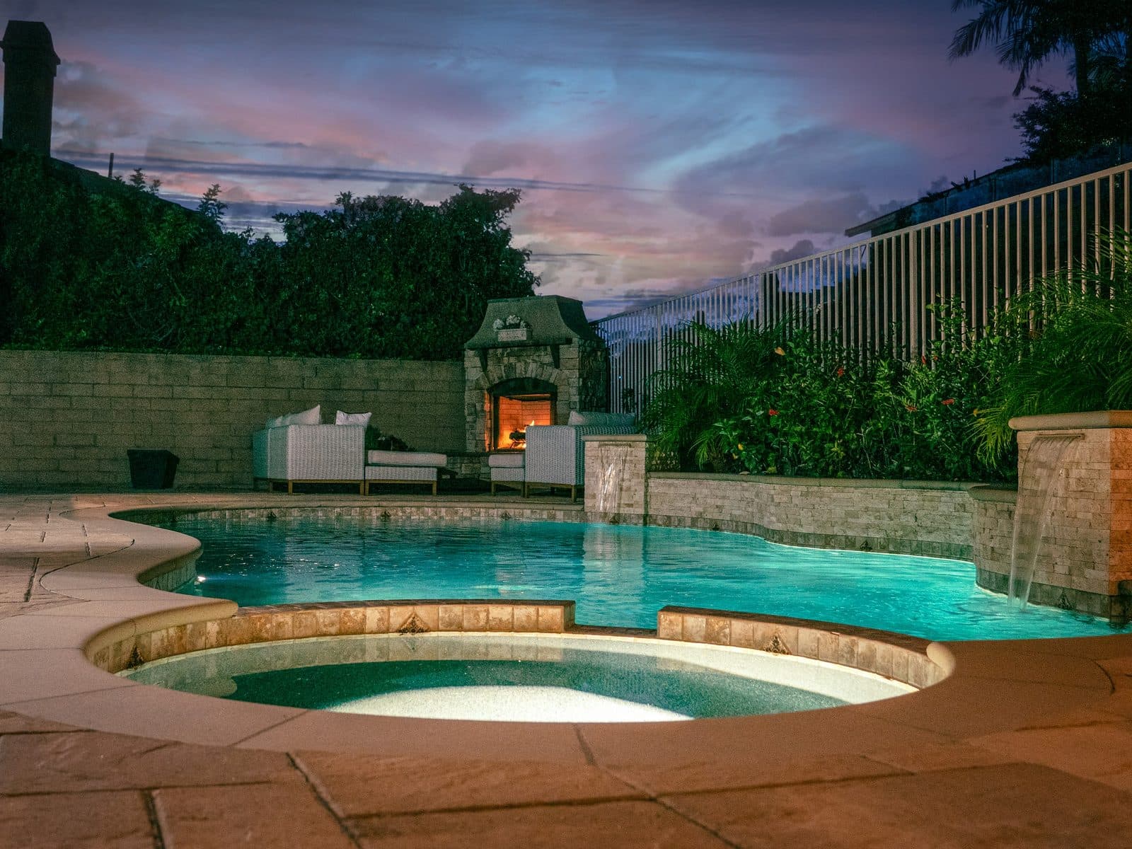 A round raised spa spilling into a pool at dusk, with a limestone wall and outdoor fireplace behind it.