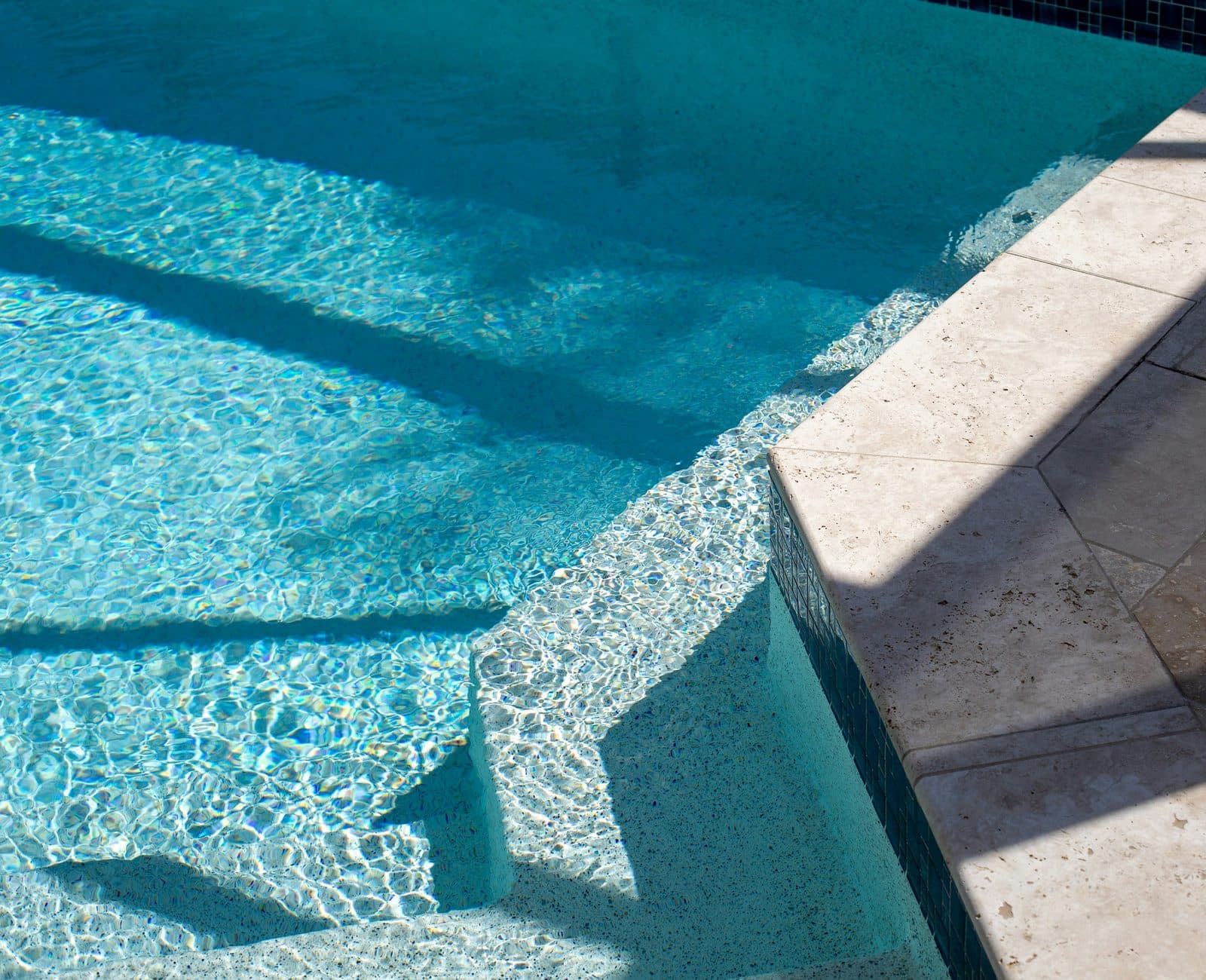 Clear pool water beside travertine coping, with hard afternoon shadows across the deck.